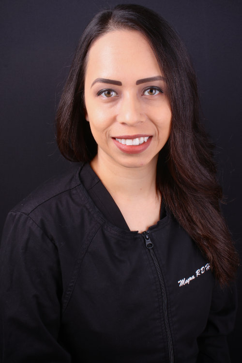 Portrait of a smiling woman with long dark hair wearing a black jacket with "megan rdh" embroidered on it against a black background.